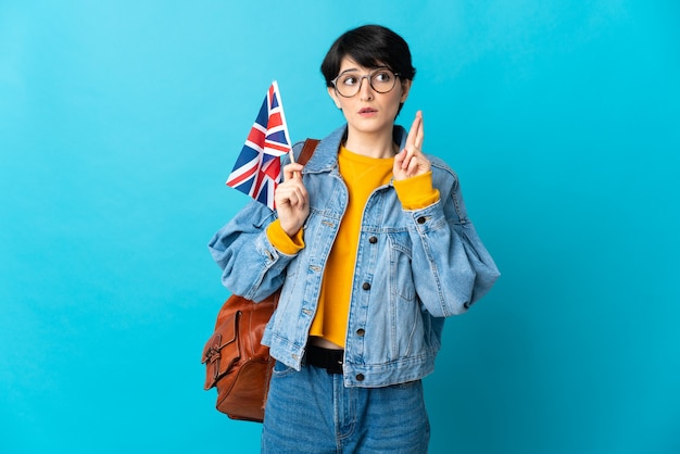 Frau mit den kurzen Haaren, die eine britische Flagge lokalisiert auf der blauen Wand mit den Fingern kreuzend halten und das Beste wünschen