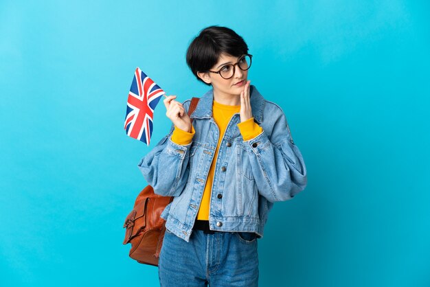 Frau mit den kurzen Haaren, die eine Flagge des Vereinigten Königreichs lokalisiert auf blauem Raum halten, der oben während des Lächelns schaut