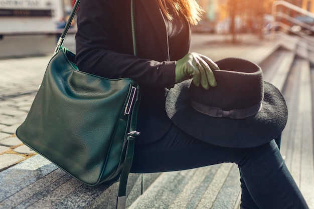 Frau mit der grünen Handtasche, die Handschuhe trägt und schwarzen Hut draußen hält