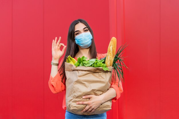 Frau mit der medizinischen maske, die eine einkaufstasche voll frisches essen hält. junge frau mit einer einkaufstasche des lebensmittels während der covid 19, coronavirus-pandemie.