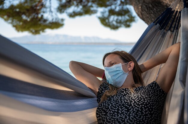 Frau mit der medizinischen Schutzmaske, die in einer Hängematte durch das Meer entspannt