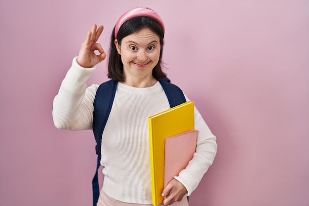 Frau mit Down-Syndrom, die einen Studentenrucksack trägt und Bücher hält, die positiv lächeln und mit der Hand und den Fingern einen erfolgreichen Ausdruck machen