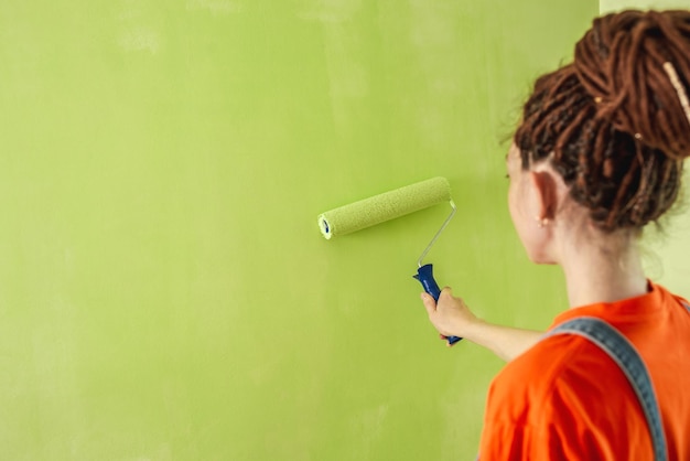 Frau mit Dreadlocks in einem orangefarbenen T-Shirt und Jeansoverall malt die Wand mit einer Walze in grüner Farbe Konzept der Reparaturrenovierung der neuen Wohnung Kopierraum