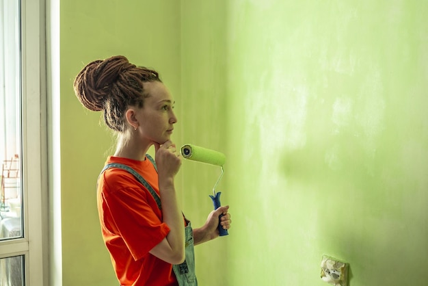 Frau mit Dreadlocks in einem orangefarbenen T-Shirt und Jeansoverall malt die Wand mit einer Walze in grüner Farbe Konzept der Reparaturrenovierung der neuen Wohnung Kopierraum