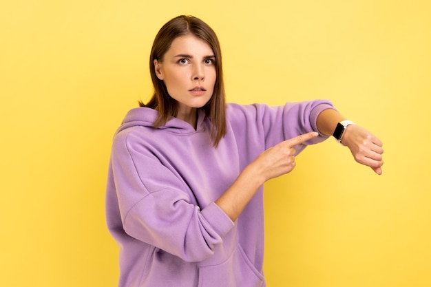 Foto frau mit dunklem haar, die auf eine armbanduhr zeigt und verärgert und unzufrieden aussieht, zeigt die uhr, um sich zu beeilen