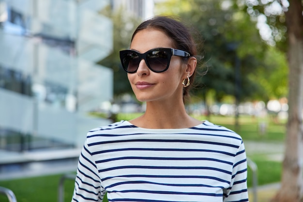 Frau mit dunklen Haaren trägt eine trendige Sonnenbrille gestreifter Pullover Spaziergänge im Freien posiert im Park Spaziergänge durch die Stadt genießt gutes Frühlingswetter