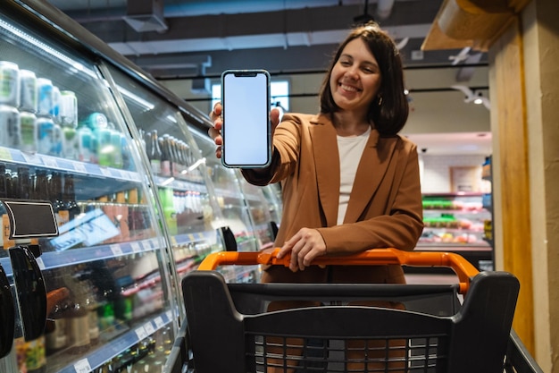 Frau mit einem Einkaufswagen, die ein Telefon mit einem weißen Bildschirm in einem Supermarkt hält