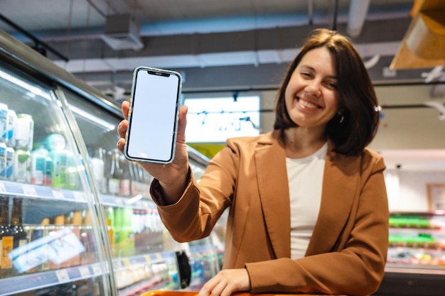 Frau mit einem Einkaufswagen, die ein Telefon mit einem weißen Bildschirm in einem Supermarkt hält