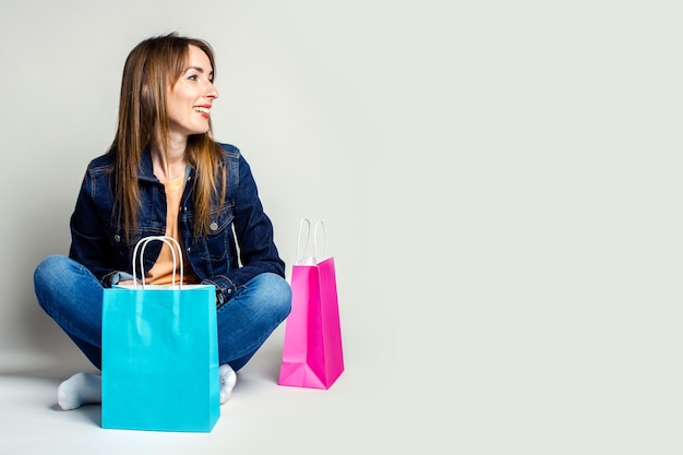 Frau mit einem Lächeln in einer Jeansjacke hält Einkaufstaschen, während sie auf dem Boden auf einem hellen Raum sitzt und zur Seite schaut. Banner.