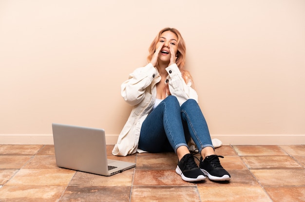 Frau mit einem Laptop, der auf dem Boden drinnen sitzt und etwas anruft und ankündigt