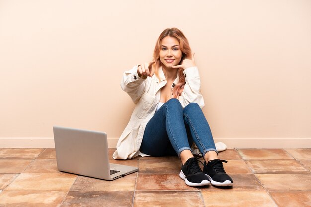 Frau mit einem Laptop, der auf dem Boden drinnen sitzt und Telefongeste macht und nach vorne zeigt