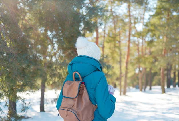 frau mit einem rucksack in einer weißen strickmütze und einer blauen jacke durch einen verschneiten winter conif