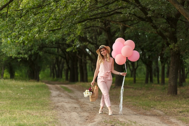 Frau mit einem Weidenkorb, einem Hut, rosa Ballons und Blumen, die auf eine Landstraße gehen