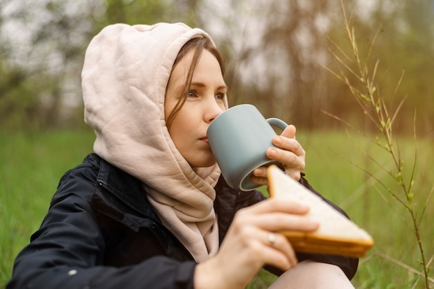 Frau mit einer Tasse im Wald, parken Sie bei kühlem Wetter auf einem grünen Hintergrund aus Laub. Entspannung in der Natur, Ruhe.