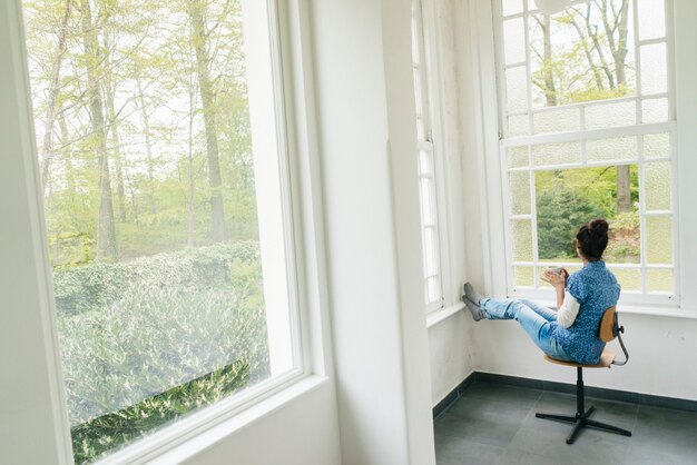 Frau mit einer Tasse Kaffee sitzt auf einem Stuhl am Fenster