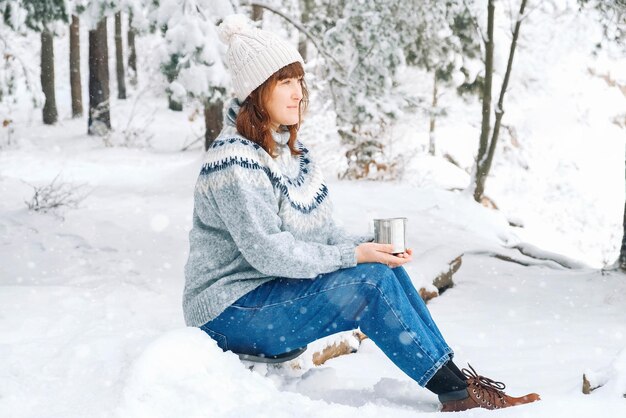 Frau mit einer Tasse Tee in ihren Händen, die in einem schneebedeckten Wald sitzt