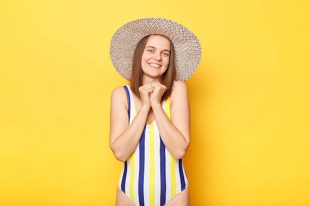 Frau mit fröhlichem Gesichtsausdruck trägt gestreiften Badeanzug und Sonnenhut, während sie isoliert auf gelbem Hintergrund steht und direkt in die Kamera blickt und einen ruhigen Tag am Strand genießt