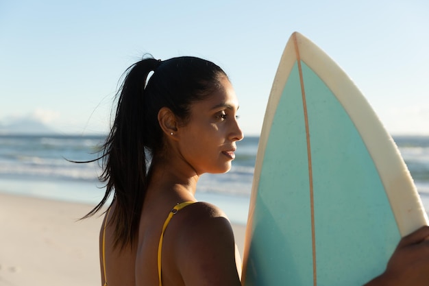 Frau mit gemischter Abstammung trägt Surfbrett am Strand. Gesunde Freizeit im Freien am Meer.