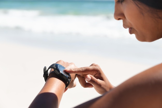 Frau mit gemischter Abstammung trainiert am Strand und überprüft ihre Smartwatch. Gesunde Freizeit im Freien am Meer.