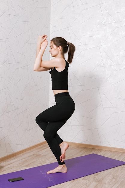 Frau mit geschlossenen Augen, die in der Adler-Pose steht und während der Yoga-Sitzung in der Wohnung balanciert