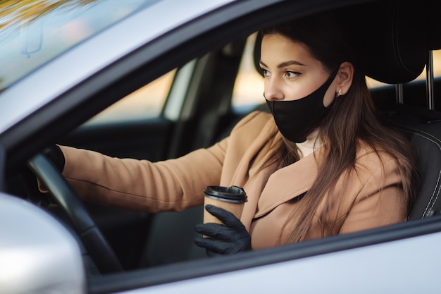 Frau mit Gesichtsmaske, die ihr Auto mit Tasse Kaffee während der Coronavirus-Pandemie fährt,