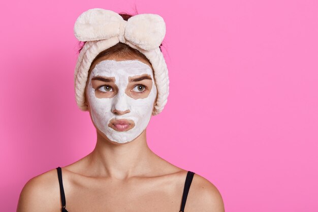 Frau mit Gesichtsmaske, die mit nachdenklichem Gesichtsausdruck beiseite schaut, lustiges Haarband habend, das über rosa Wand lokalisiert aufwirft. Kopieren Sie Platz für Werbung.