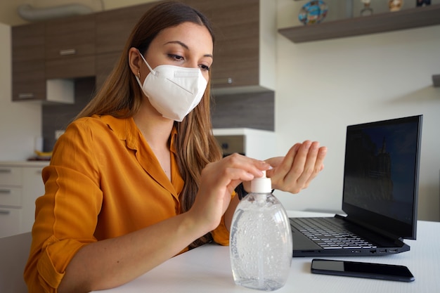 Frau mit Gesichtsmaske, die von zu Hause aus mit Laptop arbeitet und Handalkoholgel verwendet