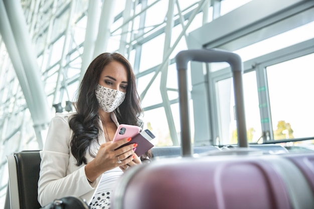 Frau mit Gesichtsmaske überprüft ihr Telefon, während sie in einer Halle auf ein Flugzeug wartet und einen Reisepass und eine Bordkarte hält.