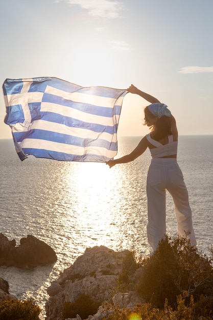 Frau mit griechischer Flagge, die den Sonnenuntergang über dem Meer betrachtet