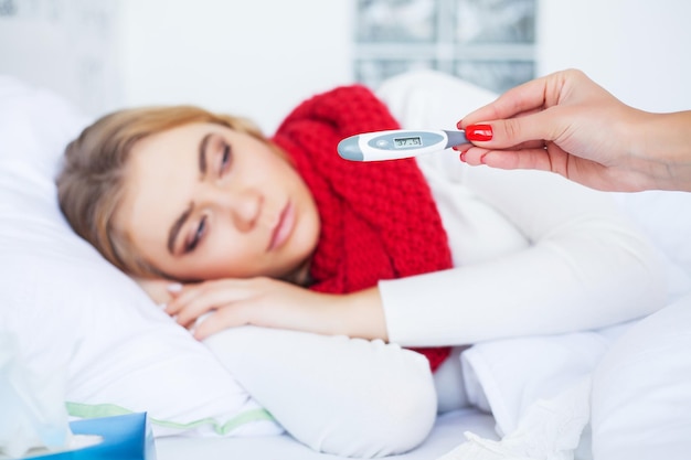 Frau mit Grippevirus liegt im Bett, sie misst ihre Temperatur mit einem Thermometer