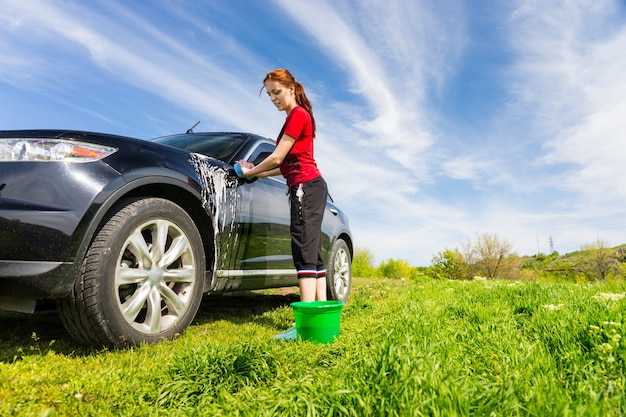 Frau mit grünem Eimer wäscht schwarzes Luxusfahrzeug im Feld mit seifigem Schwamm an einem hellen sonnigen Tag