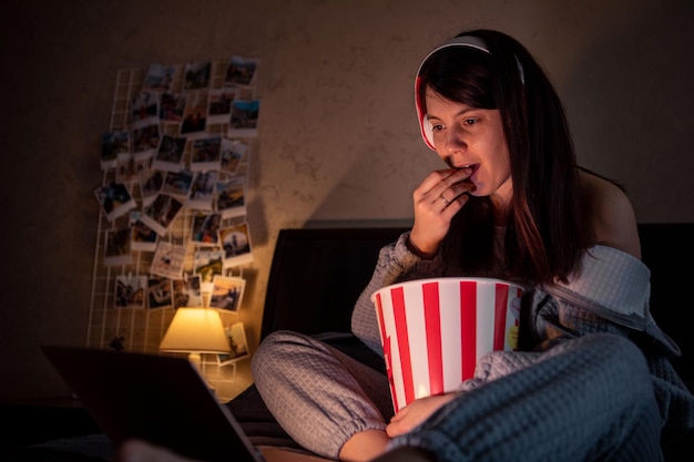 Frau mit Headset, die auf dem Bett sitzt und Popcorn isst und nachts Filme sieht