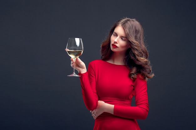 Frau mit hellem Make-up, Frisur, die rotes Kleid trägt, das mit Glas Weinstock über Dunkelheit aufwirft