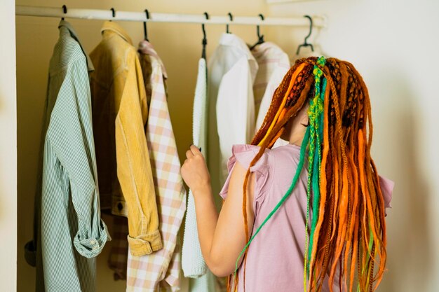 Frau mit hellen Dreadlocks, die Kleidung in der Umkleidekabine auswählt Moderne leichte Damenumkleidekabine mit verschiedenen Dingen