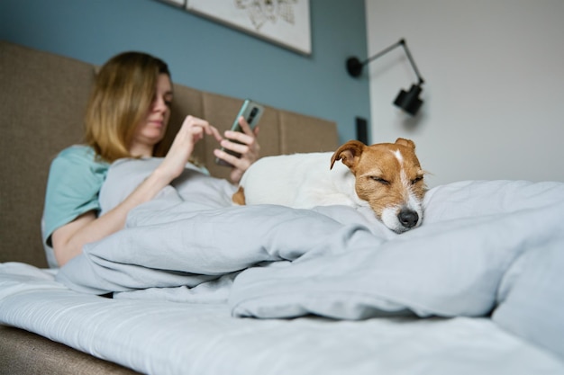 Frau mit Hund, die auf dem Bett chillt, benutzt Smartphone