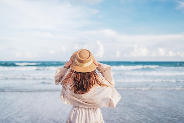 Frau mit Hut entspannt sich mit erhobenen Armen auf den Kopf und genießt den Blick auf den Sommertag am Strand