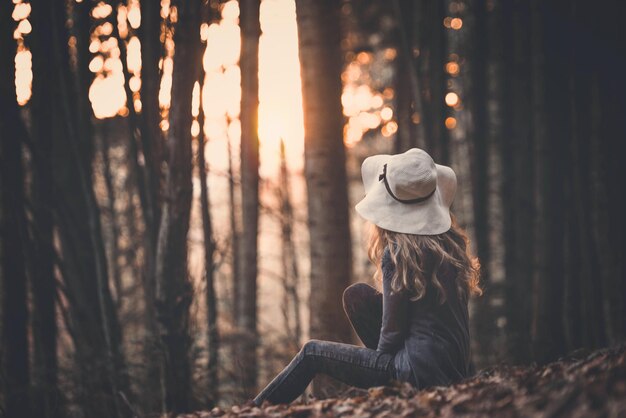 Frau mit Hut sitzt während des Sonnenuntergangs auf dem Feld gegen Bäume