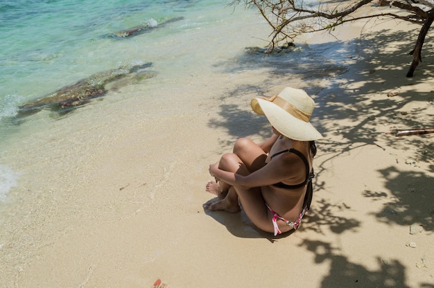 Frau mit Hut und Bikini sitzt im Schneidersitz vor dem Inselstrand in Cartagena Kolumbien