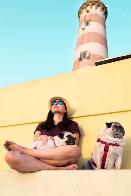 Frau mit Hut und Sonnenbrille mit ihren Hunden bei Sonnenuntergang neben dem Leuchtturm von Aveiro in Portugal