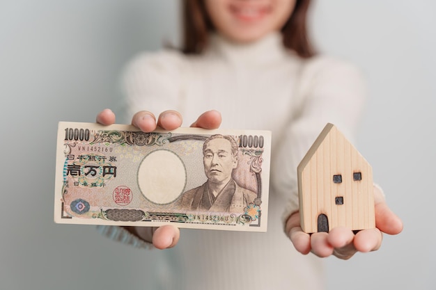 Frau mit japanischer Yen-Banknote und Hausmodell Immobilien Home Hypothek Japan Bargeld Steuerrezession Wirtschaft Inflation Investitionsfinanzierung und Sparkonzepte