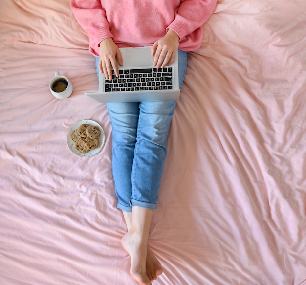 Frau mit Jeans sitzt auf dem rosa Bett mit Tasse Kaffee und arbeitet an ihrem Laptop. USA