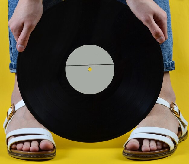 Frau mit Jeans und Sandalen hält eine Retro-Vinylplatte an einer gelben Wand