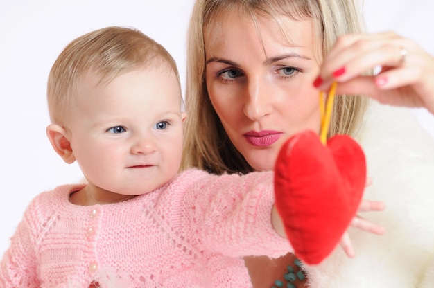 Frau mit Kind, das mit Interesse Spielzeug in Form des roten Herzens betrachtet. Baby mit grauen Augen in rosa Kleidern