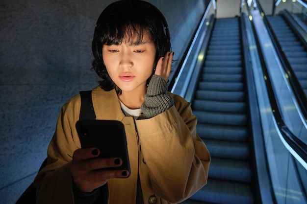 Frau mit Kopfhörern hört Musik und schaut besorgt auf den Bildschirm ihres Telefons, der die Rolltreppe in der Stadt hinunterfährt