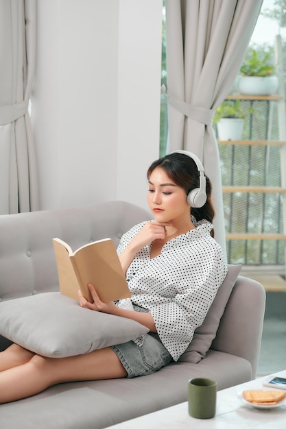 Frau mit Kopfhörern liest ein Buch im Wohnzimmer, das auf der Couch sitzt