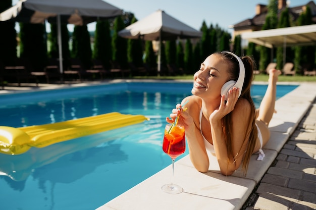 Frau mit Kopfhörern trinkt Cocktail am Pool