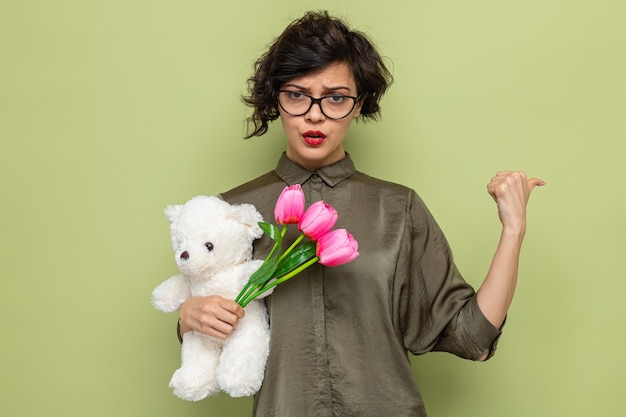 Frau mit kurzen Haaren, die einen Strauß Tulpen und einen Teddybären hält, der mit ernstem Gesicht schaut, das mit dem Daumen nach hinten zeigt