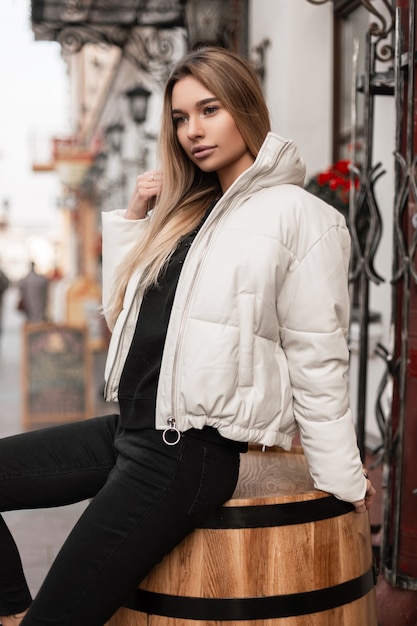 Frau mit langen blonden Haaren in einer trendigen weißen Jacke sitzt auf einem hölzernen Vintage-Fass