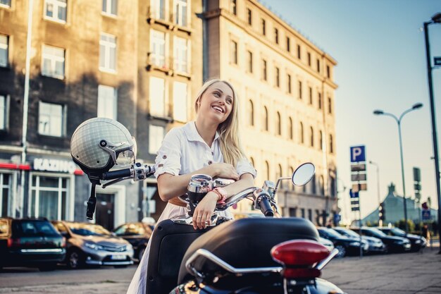 Frau mit langen blonden Haaren in weißem Kleid posiert in der Nähe eines Rollers auf der Straße in der Stadt.