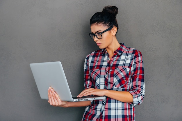 Frau mit Laptop. Schöne junge Frau, die am Laptop arbeitet, während sie vor grauem Hintergrund steht